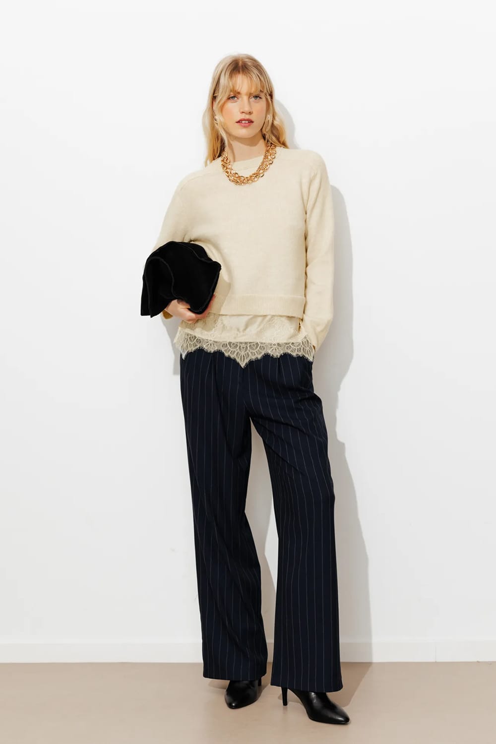 Look femme hiver 2026 avec pull écru à dentelle, pantalon large à rayures, boots noires et pochette en velours