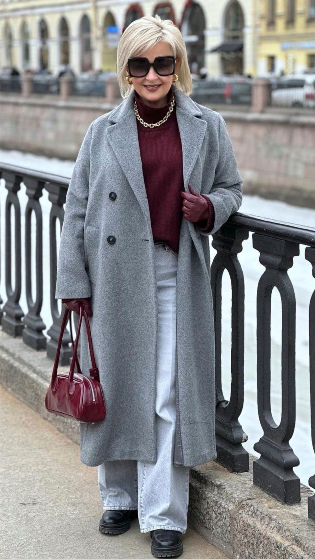 Tenue hiver femme 60 ans avec manteau long gris, pull bordeaux, jean clair, sac bordeaux et chaussures noires