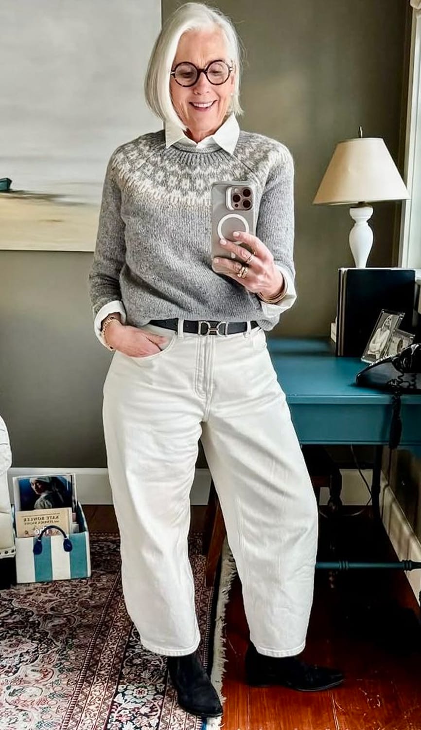 Look d’hiver moderne pour femme de 60 ans : pull jacquard gris sur chemise blanche, jean blanc taille haute et bottines noires, style casual chic.