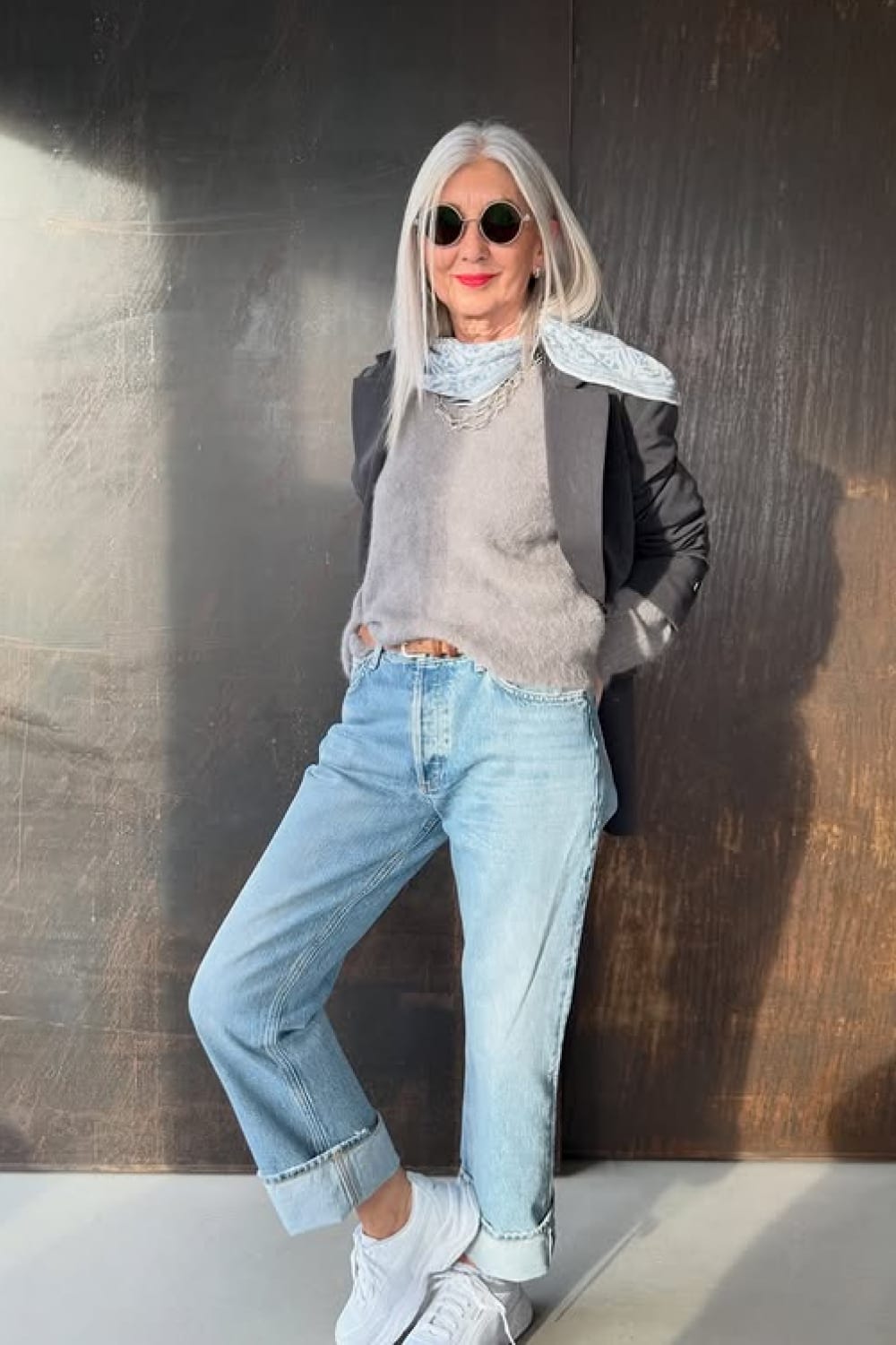 Tenue 60 ans moderne : blazer noir sur pull gris, jean clair coupe droite et baskets blanches, foulard bleu clair.
