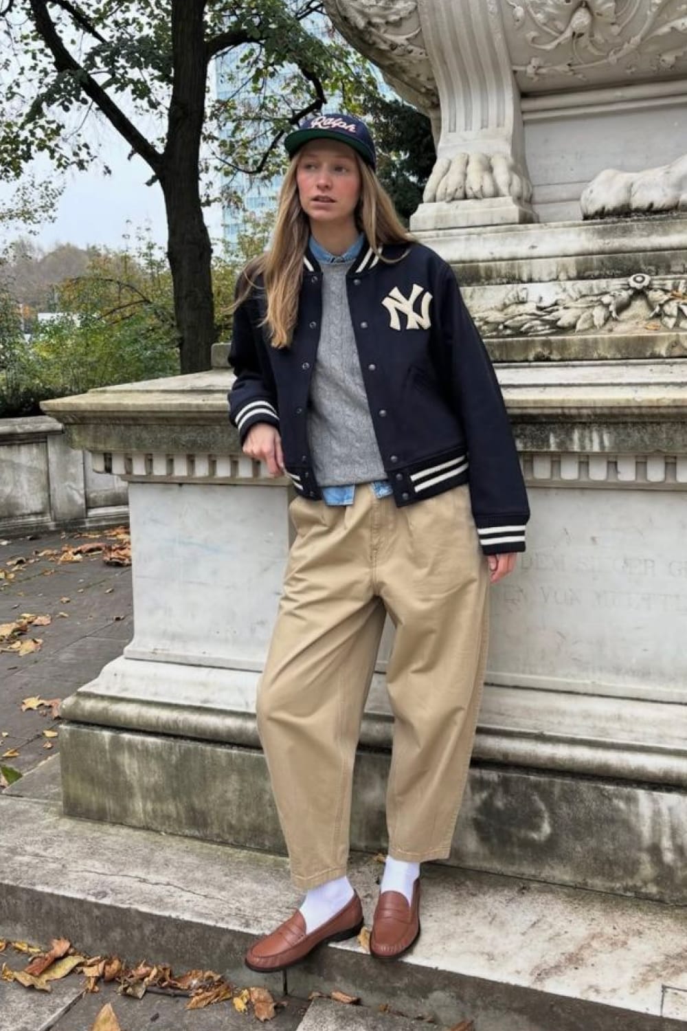 Blouson teddy varsity marine à écusson et bandes crème, porté sur sweat gris et pantalon beige, tendance printemps 2026