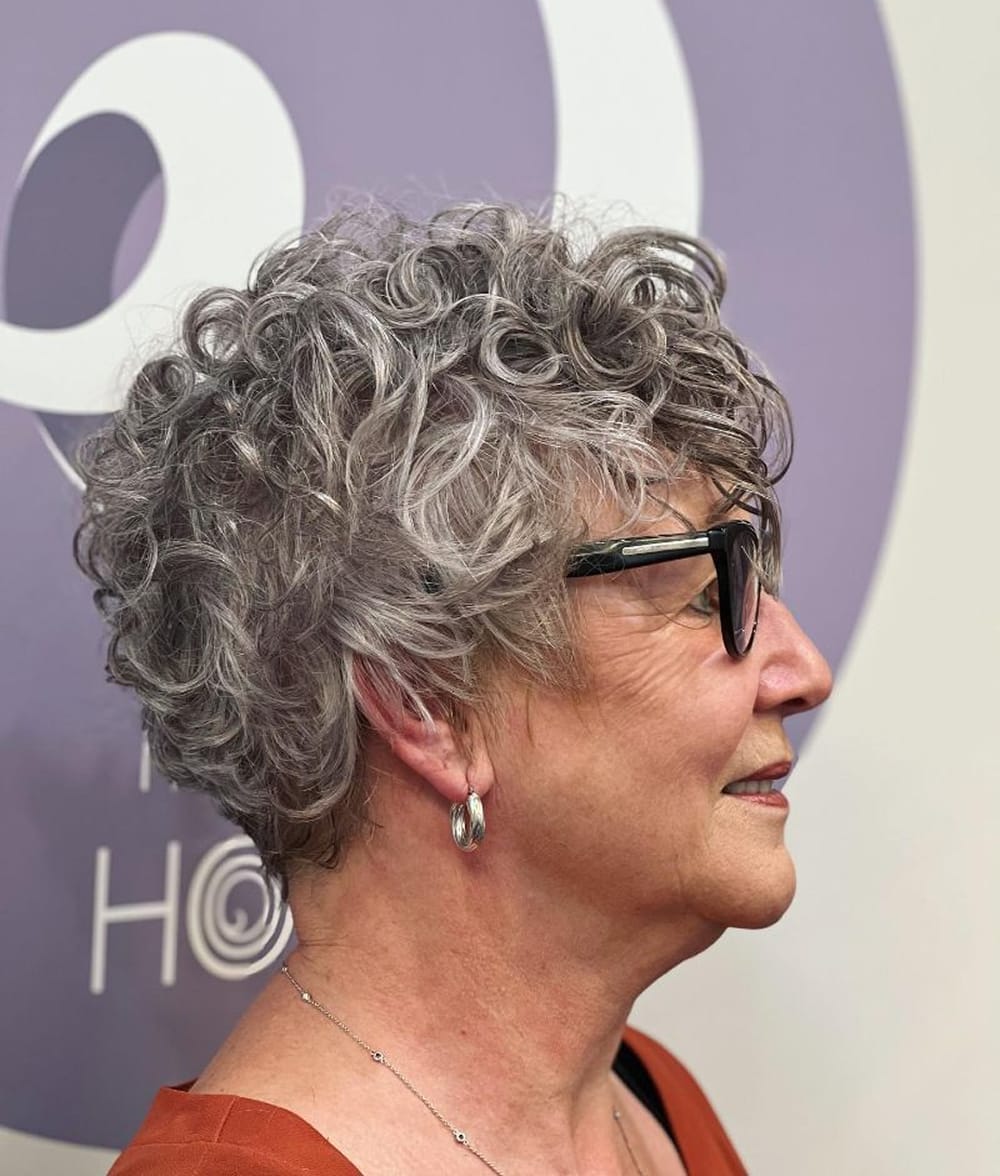 Coupe courte bouclée serrée type 3B/3C gris poivre et sel, profil femme senior avec lunettes