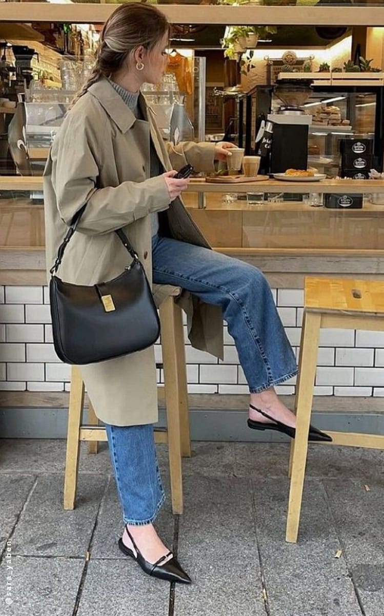 Trench beige porté avec un pull gris à col montant, un jean droit et des slingbacks noires dans un café
