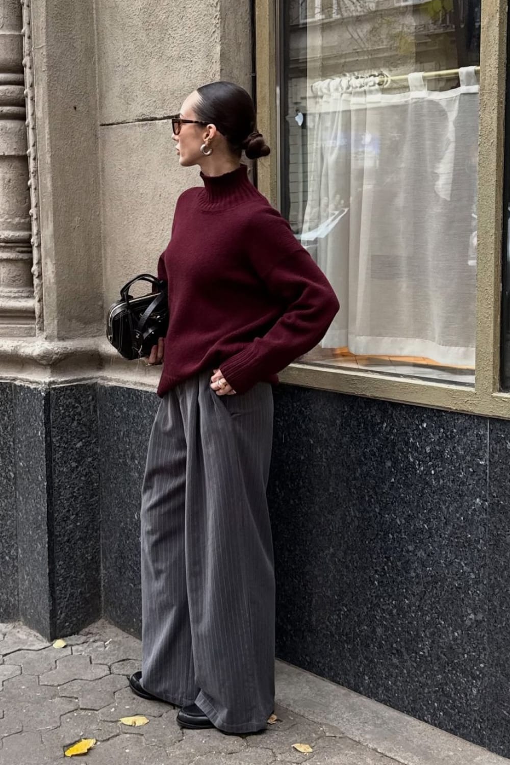 Femme portant un pantalon gris large à fines rayures avec un pull col montant bordeaux oversize, des mocassins noirs et un sac à main noir, look d'automne en ville