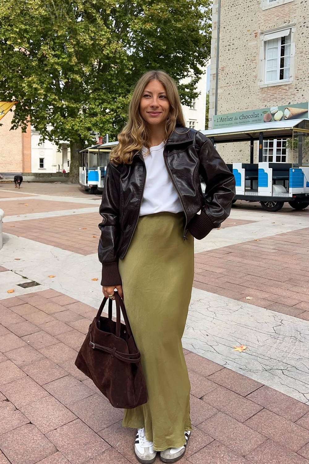 Jupe longue satinée vert olive clair avec un haut blanc, un blouson court en cuir marron chocolat, un sac en daim et des baskets blanches