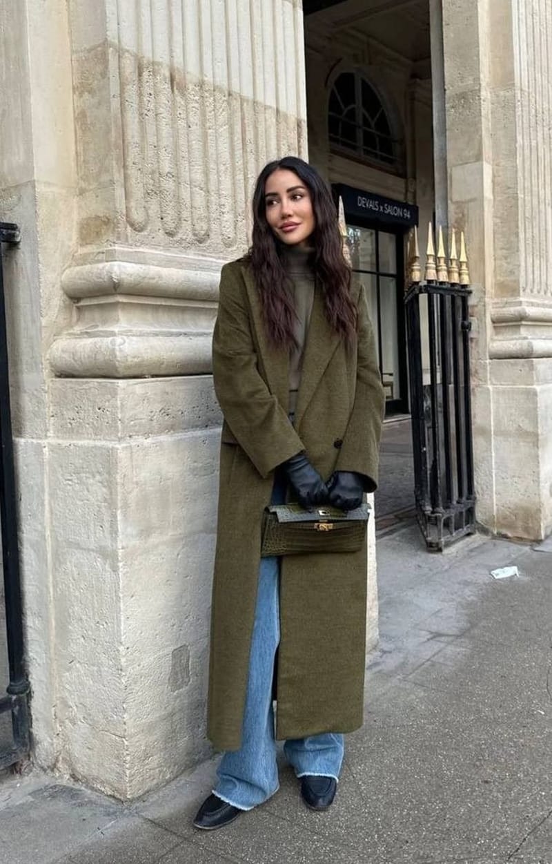 Manteau long vert olive porté ouvert sur un col roulé kaki, un jean large clair, des gants en cuir noir et un sac en croco olive