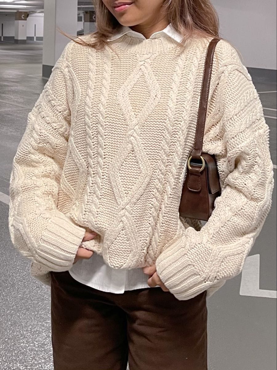 Pull en maille torsadée écru porté sur une chemise blanche avec un pantalon chocolat, look d'automne en couches