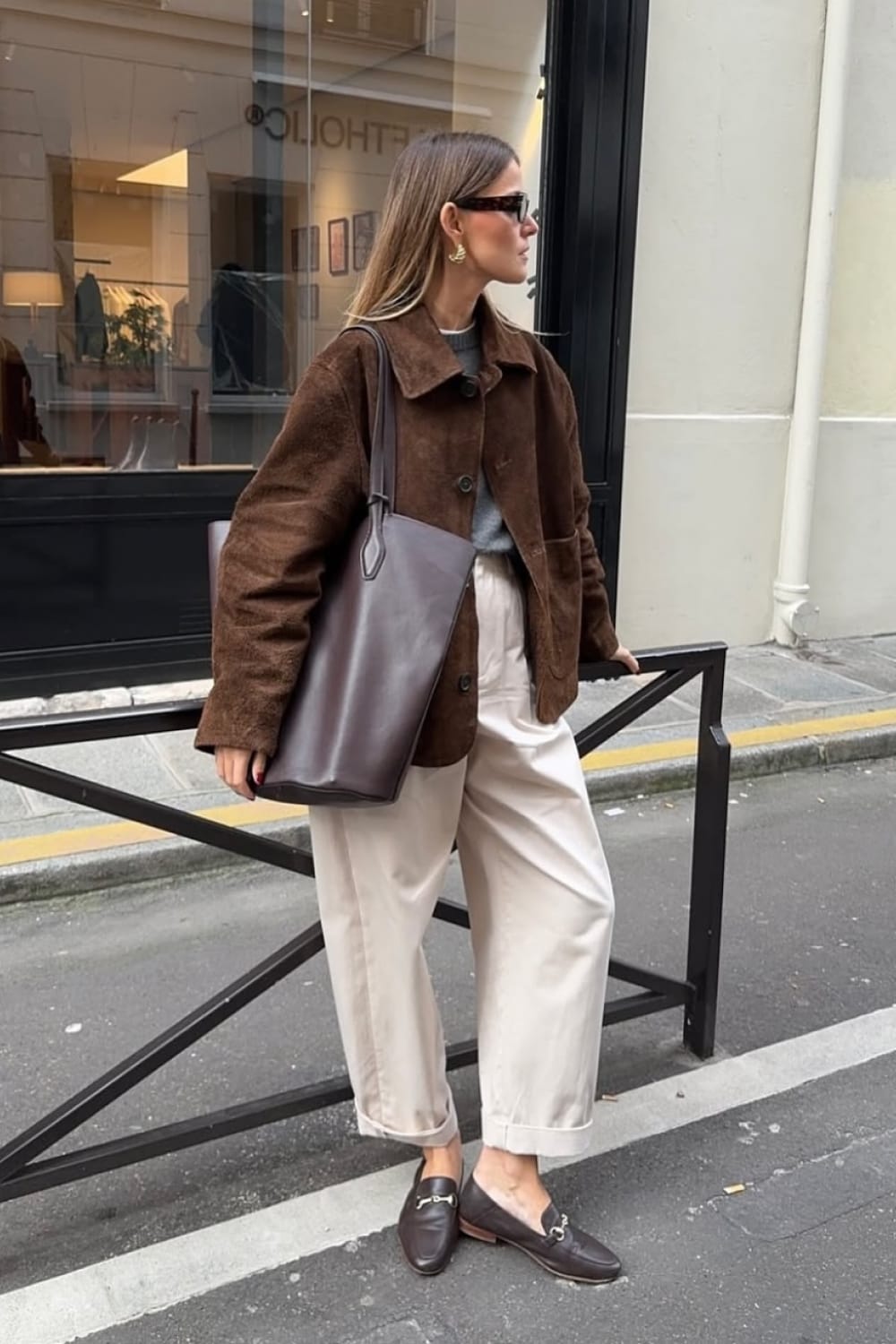 Tenue automne femme avec veste surchemise en daim marron foncé ouverte sur un pull gris, avec un pantalon à pinces écru retroussé, des mocassins en cuir brun et un grand cabas en cuir