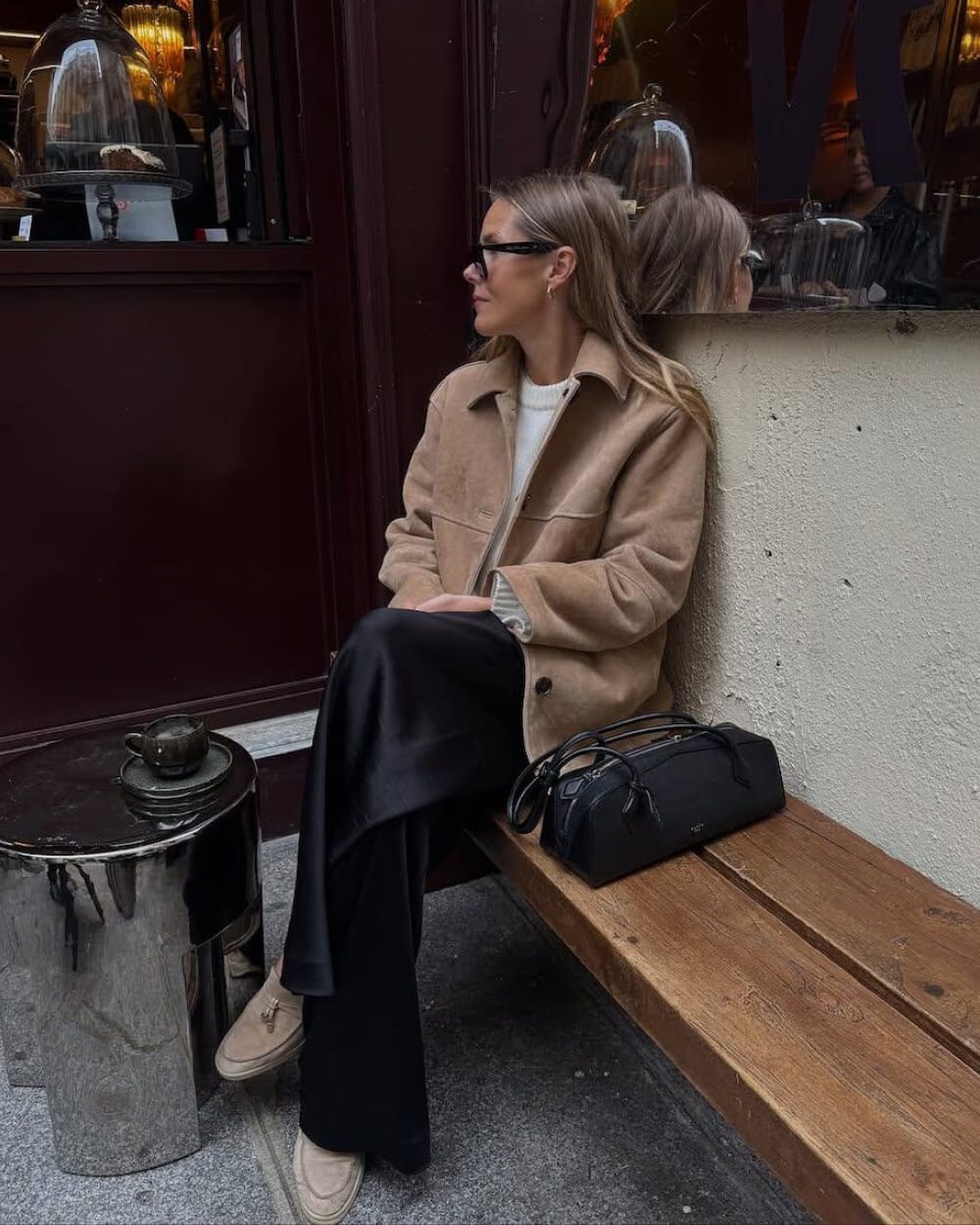 Veste en cuir velours camel portée sur un pull en grosse maille crème et un pantalon large en satin noir, avec des mocassins en daim beige et un sac noir structuré