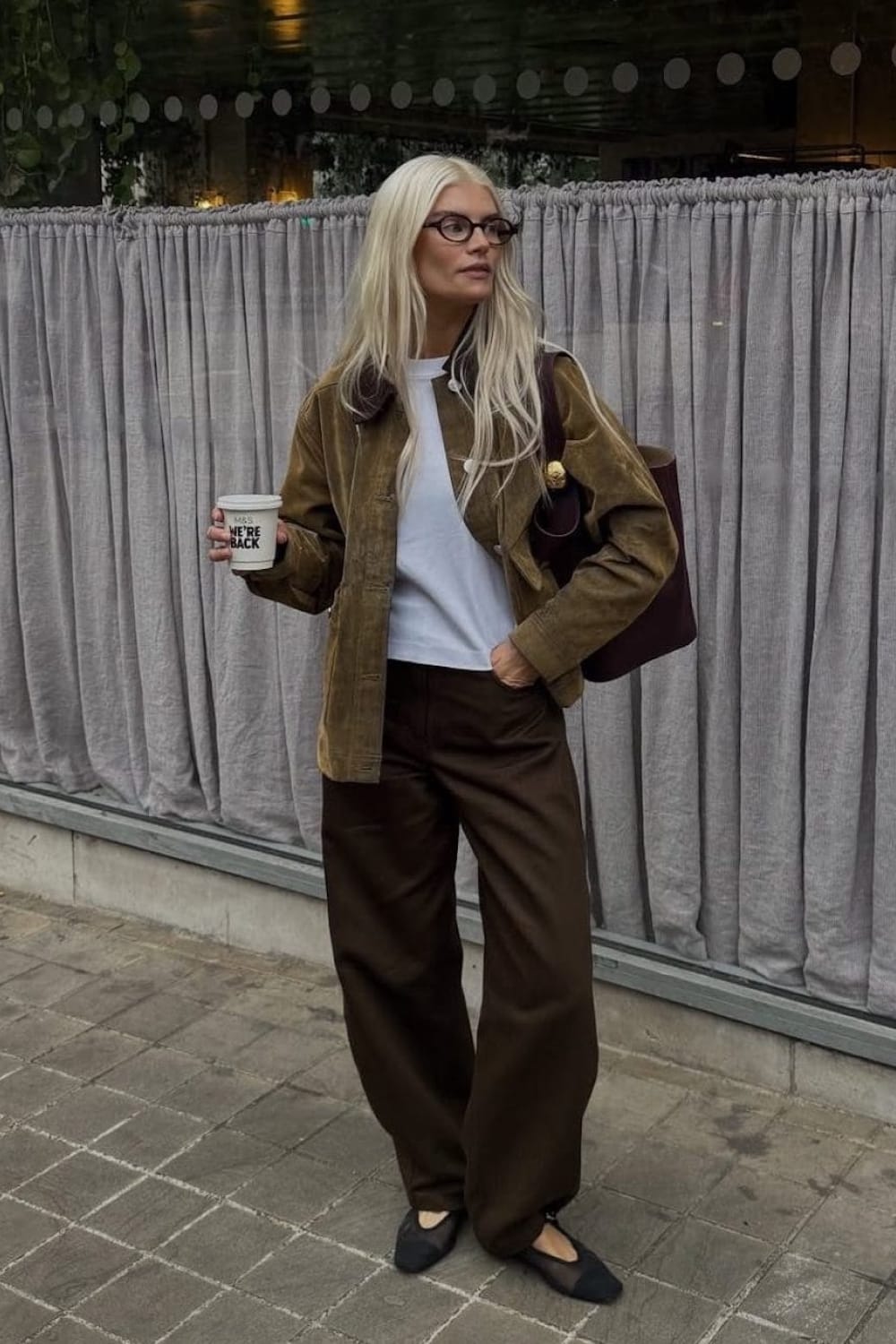 Veste en suède kaki à col marron contrasté portée avec un tee shirt blanc, un pantalon large chocolat, des ballerines en résille noire et un cabas bordeaux