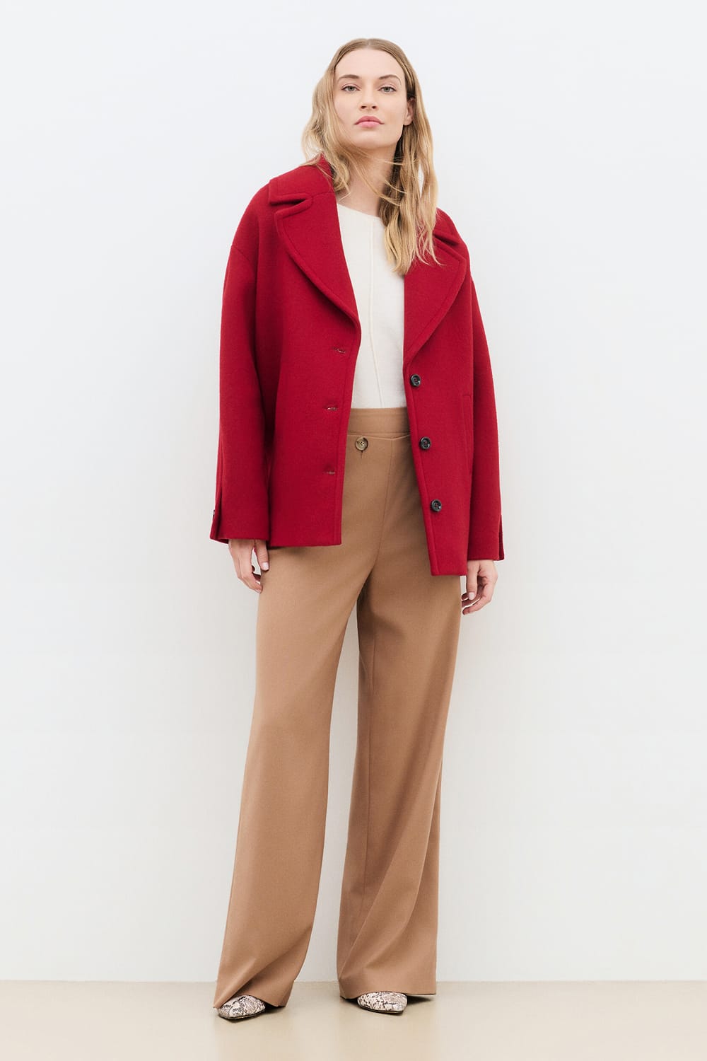 Look collection Caroll automne-hiver 2026-2027 avec manteau court rouge à larges revers, porté ouvert sur un haut écru, pantalon large camel et chaussures à bout pointu imprimées